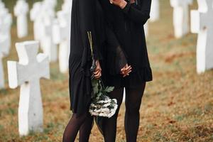 Holds flowers. Two young women in black clothes visiting cemetery with many white crosses. Conception of funeral and death photo