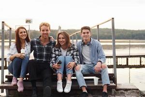 Sitting near lake and embracing each other. Group of young cheerful friends that is outdoors having fun together photo