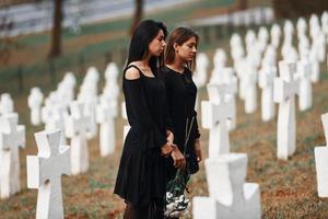 Holds flowers. Two young women in black clothes visiting cemetery with many white crosses. Conception of funeral and death photo