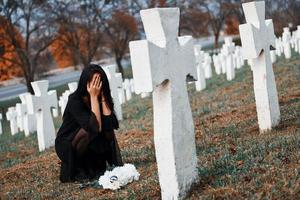 With flowers in hands. Young woman in black clothes visiting cemetery with many white crosses. Conception of funeral and death photo