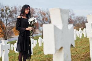 With flowers in hands. Young woman in black clothes visiting cemetery with many white crosses. Conception of funeral and death photo