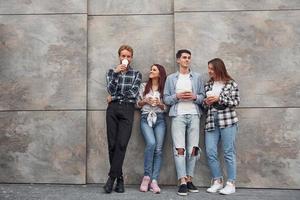 Group of young positive friends in casual clothes standing together against grey wall with cups of drink in hands photo