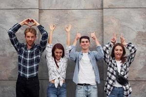 haciendo diferentes gestos. grupo de jóvenes amigos positivos en ropa casual de pie juntos contra la pared gris foto