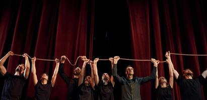 sosteniendo la cuerda en las manos por encima de la cabeza. grupo de actores con ropa de color oscuro ensayando en el teatro foto