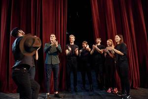 de pie contra las cortinas rojas. grupo de actores con ropa de color oscuro ensayando en el teatro foto