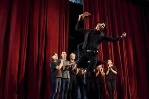 de pie contra las cortinas rojas. grupo de actores con ropa de color oscuro ensayando en el teatro foto