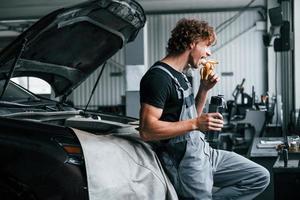 Takes a break. Eats banana and drinks water. Adult man in grey colored uniform works in the automobile salon photo