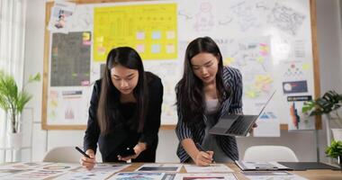 dos mujeres asiáticas revisando la tarea en el escritorio del lugar de trabajo mientras están en la oficina. una mujer sosteniendo una computadora portátil y escribiendo en papel, una mujer usando un teléfono inteligente y escribiendo notas en papeles adhesivos coloridos. video