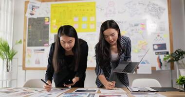 Two asian women checking to task on workplace desk while standing in office. One woman holding laptop and writing on paper, One woman using smartphone and writing notes on colorful sticky papers. video