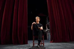 Positive young man sitting on the chair between the curtains on the stage of theater photo