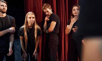 de pie contra las cortinas rojas. grupo de actores con ropa de color oscuro ensayando en el teatro foto