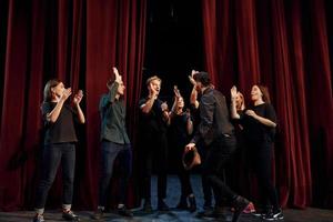 de pie contra las cortinas rojas. grupo de actores con ropa de color oscuro ensayando en el teatro foto