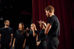 de pie contra las cortinas rojas. grupo de actores con ropa de color oscuro ensayando en el teatro foto