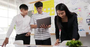 dois homens asiáticos e uma mulher verificando a tarefa na mesa do local de trabalho enquanto estão no cargo. um homem segurando laptop e escrevendo no papel, um homem escrevendo notas em papéis adesivos, mulher escrevendo no papel. video