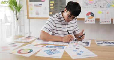 Side view of Asian man glasses checking to task on workplace desk while sitting in office. Male holding smartphone and writing on paper. Man using mobile phone. video