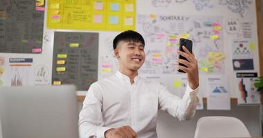 retrato do empresário atraente olhar no smartphone no local de trabalho enquanto está sentado na mesa do local de trabalho. homem sorridente usando celular na sala do escritório. homem alegre sorrindo. video