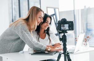 Camera on tripod is recording. Two young female freelancers working indoors in the office with laptop photo