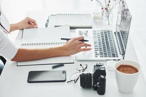 Using laptop and notepad. Young female freelancer working indoors in the office at daytime photo