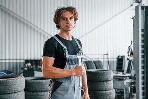 Holds wrench in hand. Adult man in grey colored uniform works in the automobile salon photo