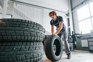 Rolls the tire. Adult man in grey colored uniform works in the automobile salon photo