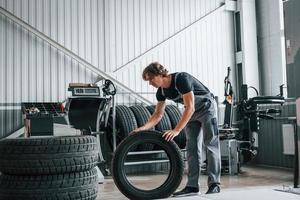 Rolls the tire. Adult man in grey colored uniform works in the automobile salon photo