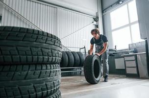 Rolls the tire. Adult man in grey colored uniform works in the automobile salon photo