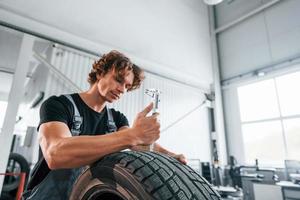 Works with tire. Adult man in grey colored uniform is in the automobile salon photo