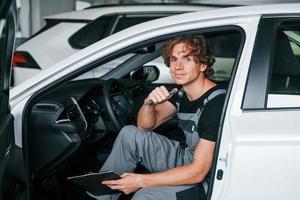 With notepad in hands. Adult man in grey colored uniform works in the automobile salon photo