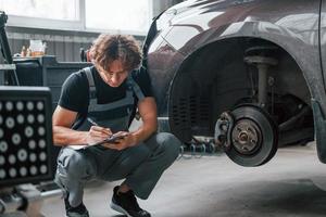 Writing in notepad. Adult man in grey colored uniform works in the automobile salon photo