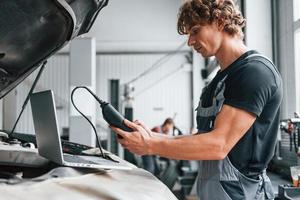 Special tool for testing characteristics. Adult man in grey colored uniform works in the automobile salon photo