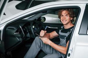 With notepad in hands. Adult man in grey colored uniform works in the automobile salon photo