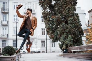 tiene sombrero y posando. modelo masculino joven en ropa de moda está al aire libre en la ciudad durante el día foto