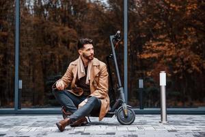 Against building and with electric scooter. Young male model in fashionable clothes is outdoors in the city at daytime photo