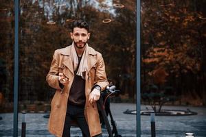 contra edificio y con patinete eléctrico. modelo masculino joven en ropa de moda está al aire libre en la ciudad durante el día foto