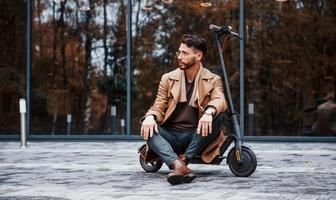 Sits on electric scooter. Young male model in fashionable clothes is outdoors in the city at daytime photo