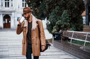 Holds cup of drink. Young male model in fashionable clothes is outdoors in the city at daytime photo