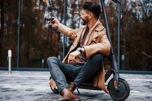 se sienta en scooter eléctrico. modelo masculino joven en ropa de moda está al aire libre en la ciudad durante el día foto