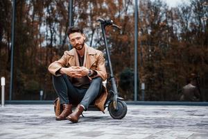 se sienta en scooter eléctrico. modelo masculino joven en ropa de moda está al aire libre en la ciudad durante el día foto