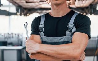 Close up view of guy that holds wrench. Adult man in grey colored uniform works in the automobile salon photo