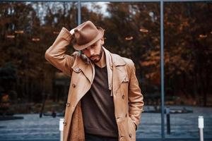 Standing against building with glass doors. Young male model in fashionable clothes is outdoors in the city at daytime photo