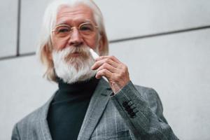 Senior businessman in formal clothes, with grey hair and beard is outdoors smokes electronic cigarette photo