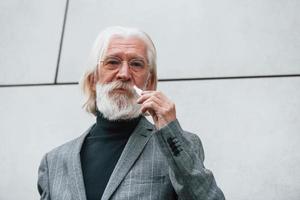 hombre de negocios senior en ropa formal, con cabello gris y barba está al aire libre fuma cigarrillo electrónico foto