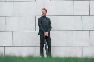 Young man in formal clothes with umbrella standing near wall outdoors with umbrella photo