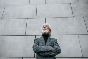 Senior businessman in formal clothes, with grey hair and beard is outdoors stands against the wall photo