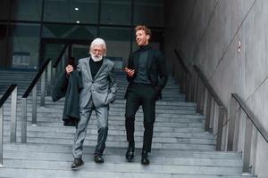 Walking on the stairs. Young guy with senior man in elegant clothes is outdoors together. Conception of business photo