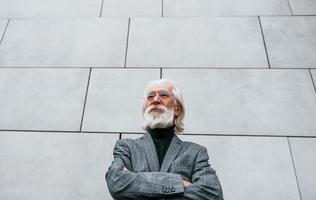 Senior businessman in formal clothes, with grey hair and beard is outdoors stands against the wall photo