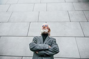 Senior businessman in formal clothes, with grey hair and beard is outdoors stands against the wall photo