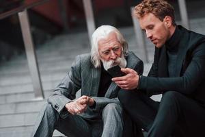 Sitting with phone. Young guy with senior man in elegant clothes is outdoors together. Conception of business photo
