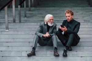 Sitting with phone. Young guy with senior man in elegant clothes is outdoors together. Conception of business photo