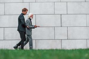 Walking on grass near wall. Young guy with senior man in elegant clothes is outdoors together. Conception of business photo